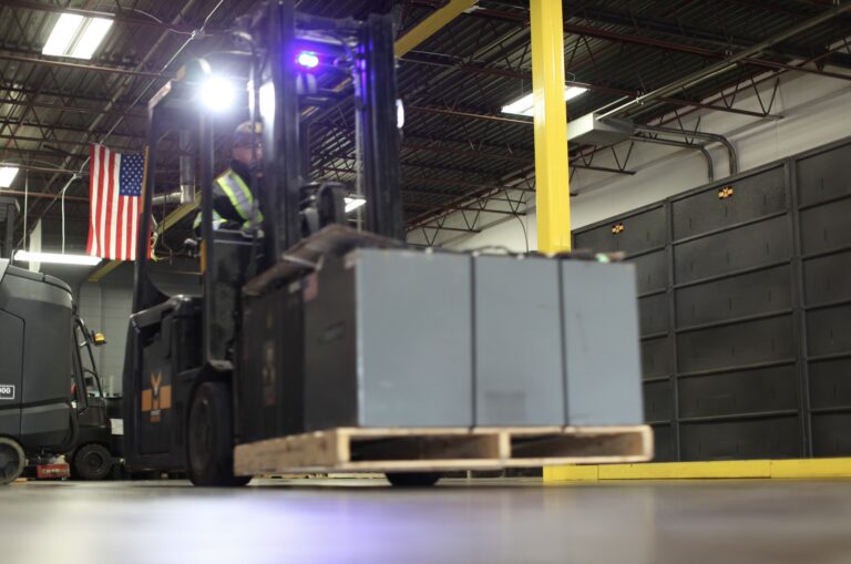 Worker positioning a large battery on forklift in store room, preparing for recycling