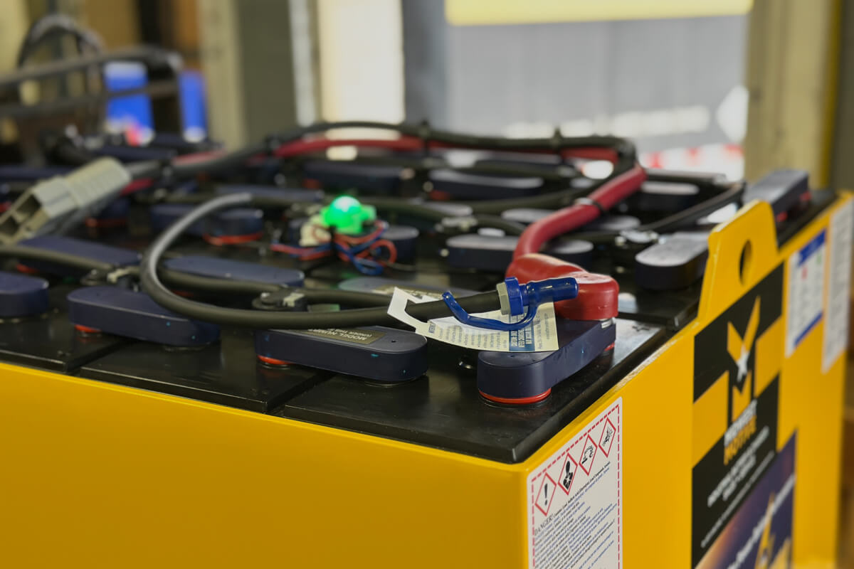 A close-up, high-angle shot of a large industrial lead-acid battery in a yellow steel case. A battery watering system with black tubing and blue connectors is visible across the top of the cells. A small green LED indicator light is glowing, and thick red and black power cables are routed along the top.